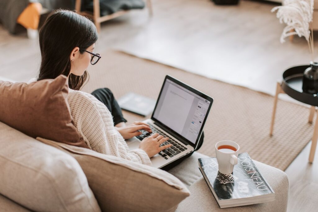 en kvinna i mörkt hår och glasögon sitter lutad mot en soffa och skriver en bloggpost om SEO på ett dokument i en laptop. Hon har en kopp med kaffe på en bok bredvid.
